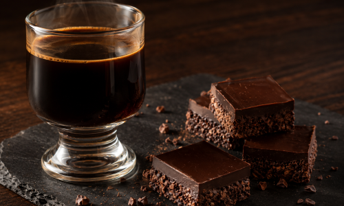 Harmonização de Café Especial com Chocolate, Quadrados de chocolate escuro bean to bar ao lado de uma taça de vidro com café especial fumegante sobre mesa de madeira.